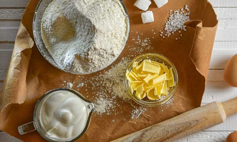 Ingredientes basicos da confeitaria, como farinhas, manteiga, cremes e leite dispostos em uma mesa de madeira.