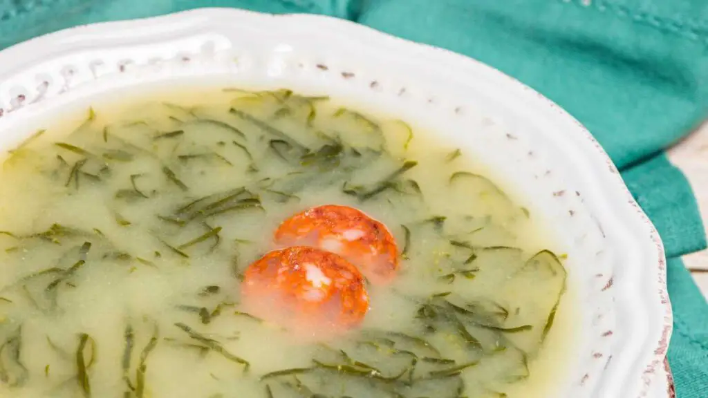 Um prato branco contendo caldo verde português. A sopa possui uma base cremosa e esverdeada, com tiras finas de couve espalhadas por toda a superfície. Fatias de chouriço flutuam no caldo, adicionando um toque de cor e sabor. O prato é finalizado com um fio de azeite de oliva, destacando a simplicidade e a riqueza dos ingredientes tradicionais.