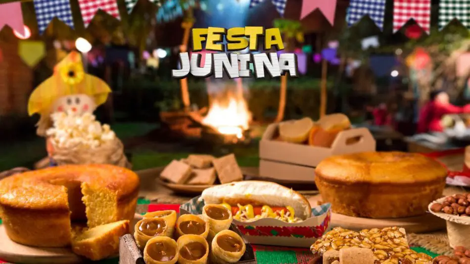 Mesa posta com comidas de festa junina e julina, mesa completa com bolo, milho cozido, bolo de milho, paçoca, caldo verde e outros. Ao fundo tem uma fogueira e bandeirolas que decoram o ambiente.