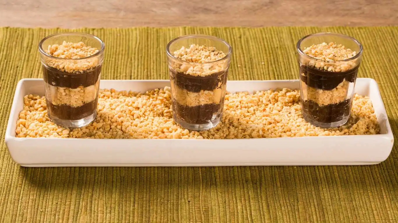 Vários brigadeiros de copinho dispostos em uma bandeja. Cada copinho é transparente, permitindo ver a textura cremosa do brigadeiro dentro. Os doces estão decorados com paçoca de amendoim no topo, e entre as camadas,. A apresentação é elegante e colorida, perfeita para festas e eventos especiais.