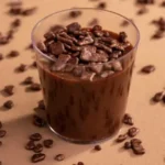 Brigadeiro de copinho disposto em uma mesa. Cada copinho é transparente, permitindo ver a textura cremosa do brigadeiro dentro. Os doces estão decorados com granulados de chocolate no topo.