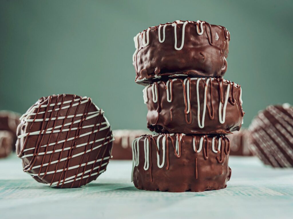 Alfajores recheados com doce de leite, cobertos com chocolate e polvilhados com açúcar de confeiteiro, dispostos em uma mesa. Os doces estão prontos para vender.