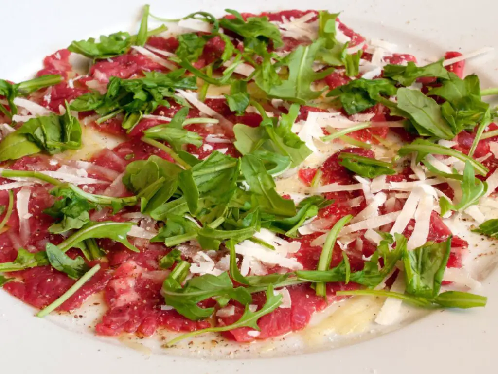 Carpaccio de carne finamente fatiado, servido em um prato com rúcula, queijo parmesão ralado e um fio de azeite.