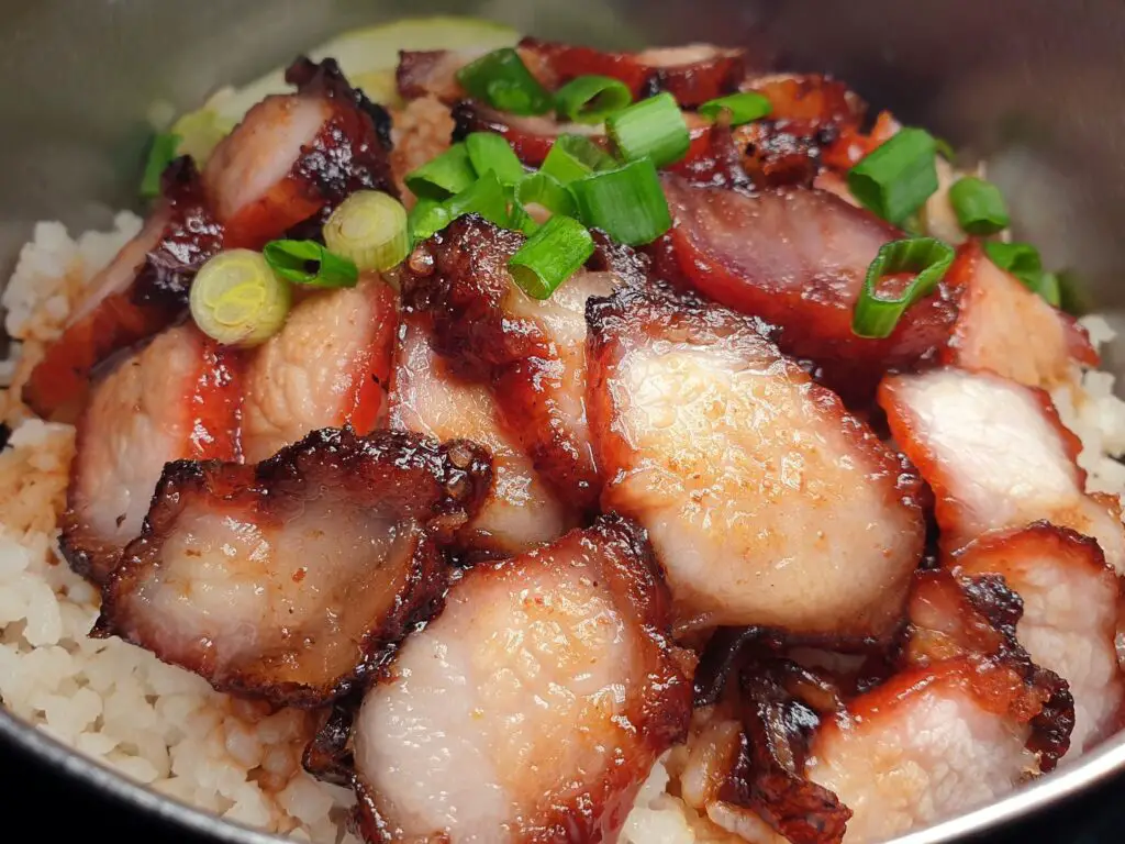 Fatias de Char Siu, carne de porco assada ao estilo chinês, com um brilho caramelizado, servidas em um prato com gergelim e cebolinha.