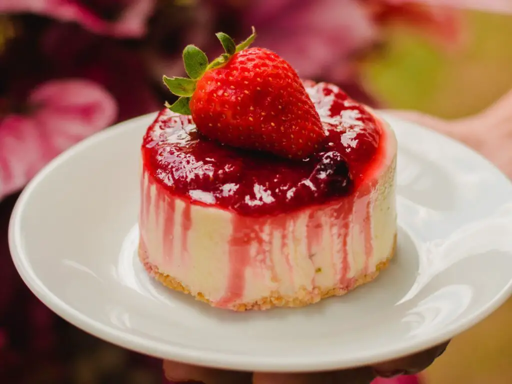 Cheesecake clássico com cobertura de frutas vermelhas, servido em um prato branco com uma base dourada de biscoito.