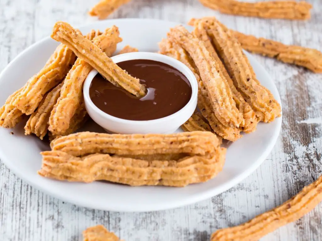 Churros quentes polvilhados com açúcar e canela, acompanhados de um recipiente com chocolate derretido.