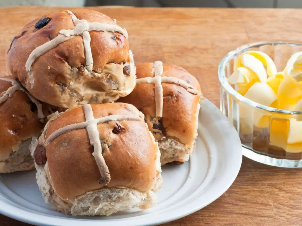 Hot Cross Buns dourados, decorados com cruzes de glacê branco, dispostos sobre uma superfície de madeira.