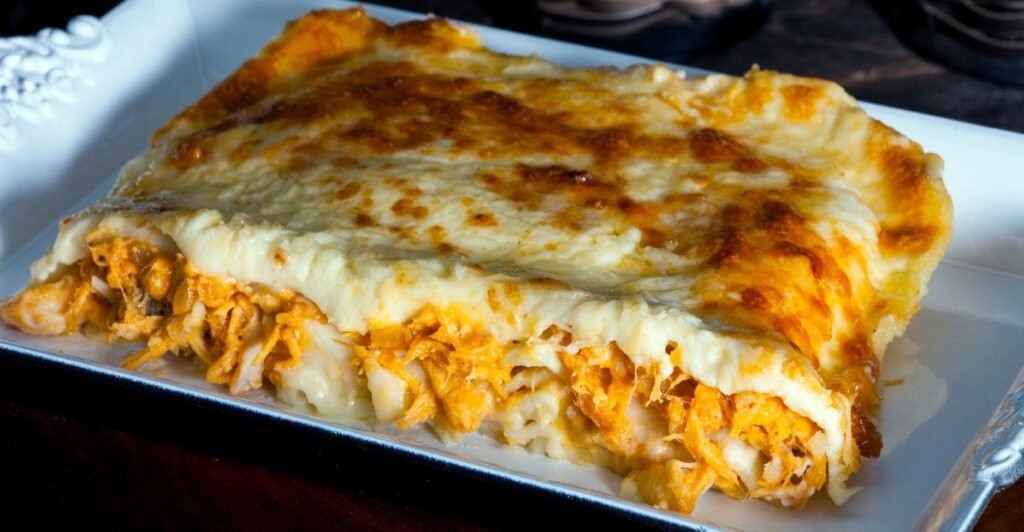 Fatia de lasanha de frango com catupiry, queijo cremoso derretido e molho ao redor, servida em prato branco.