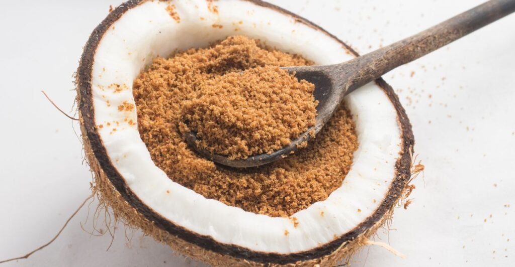 Açúcar de coco granulado em uma tigela feita da casca do coco, ao lado de um coco fresco.