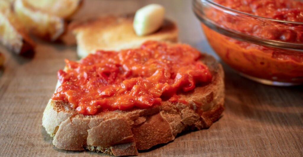 Pote de ajvar vermelho caseiro ao lado de fatias de pão rústico e pimentões assados.