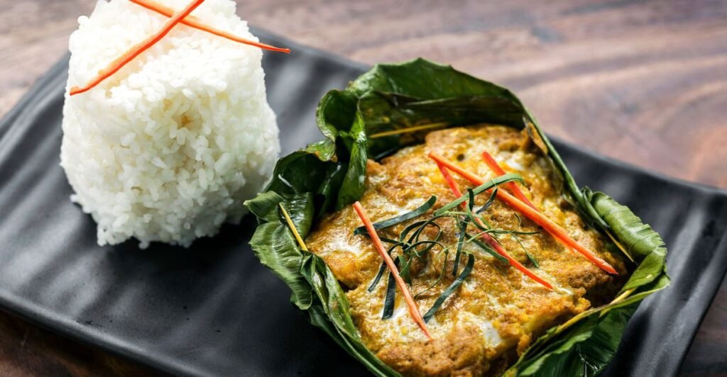 Amok trey servido em folha de bananeira com arroz branco ao lado, decorado com tiras de pimenta e folhas cítricas.