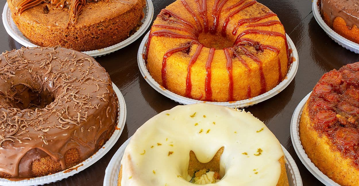 diversos bolos caseiros frescos: bolo de cenoura com chocolate, fubá cremoso, laranja e chocolate, prontos para vender com embalagens e etiquetas de marca.