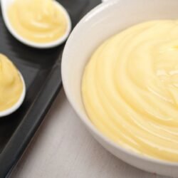 Ceramica branca cheia de brigadeiro de colher branco, cremoso e brilhante sendo exposto junto de tres colheres cheias do creme.