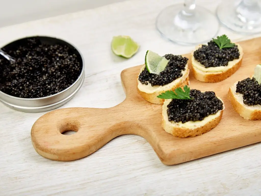 Caviar preto servido em uma pequena tigela de cristal, acompanhado por torradas finas e limão.