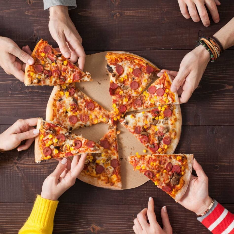 Varias pessoas pegando um pedaço de pizza em uma mesa para comemorar o dia da pizza.