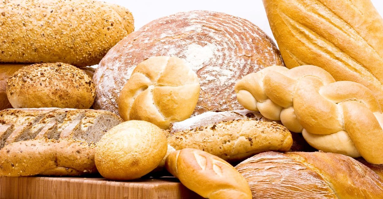 Mesa rústica com diversos tipos de pães artesanais — pães de fermentação natural, baguetes, brioches, pães integrais e pães de forma — dispostos de forma apetitosa, simbolizando o Dia Mundial do Pão.