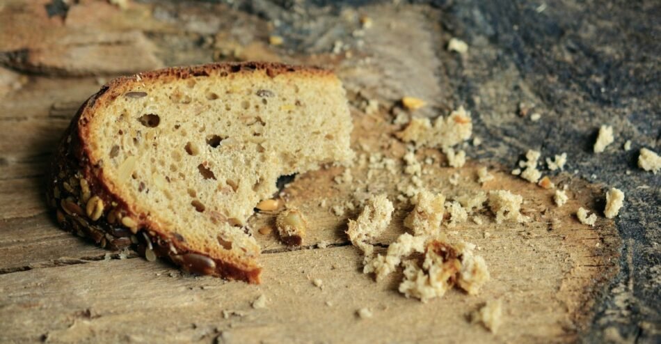 Mãos esmigalhando pedaços de pão amanhecido sobre uma tigela de vidro.