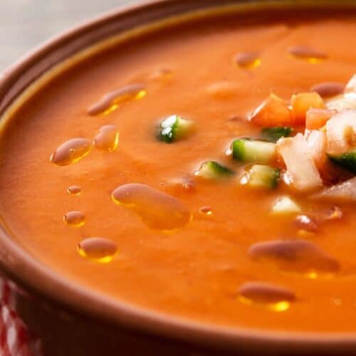 Tigela de gaspacho andaluz servido com cubinhos de pepino e pão artesanal ao lado.