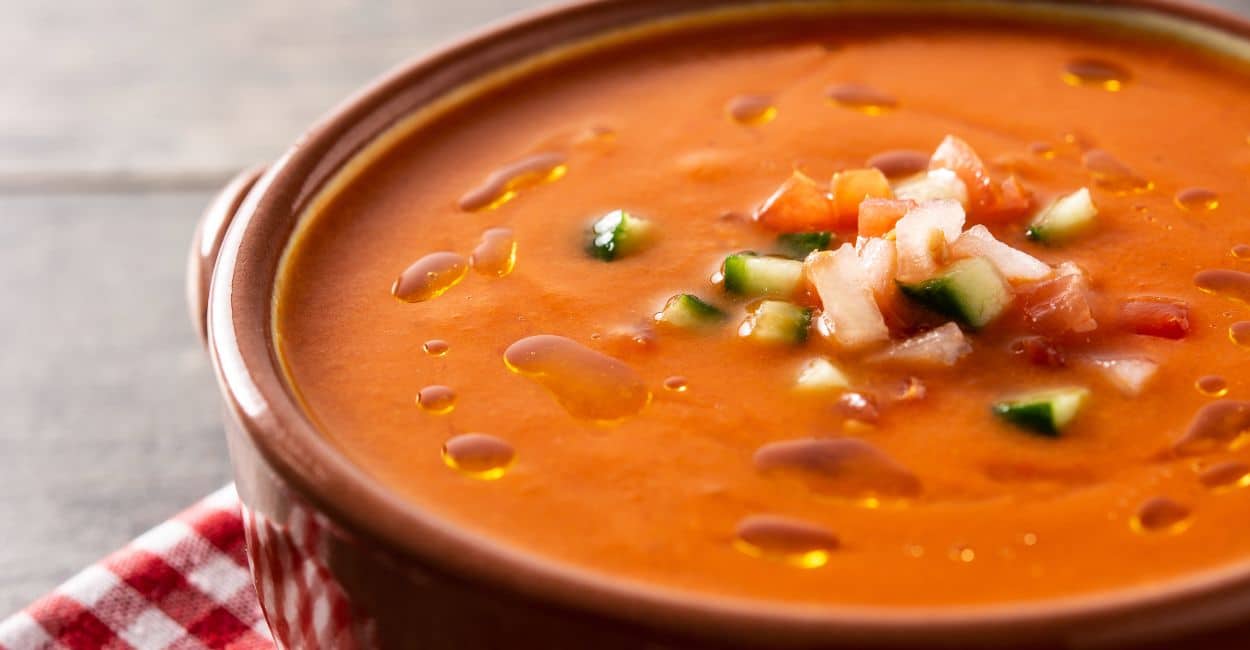 Tigela de gaspacho andaluz servido com cubinhos de pepino e pão artesanal ao lado.
