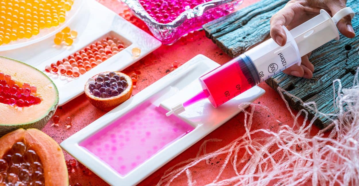 Seringa aplicando líquido colorido em gotas para formar esferas de gastronomia molecular sobre superfície branca.