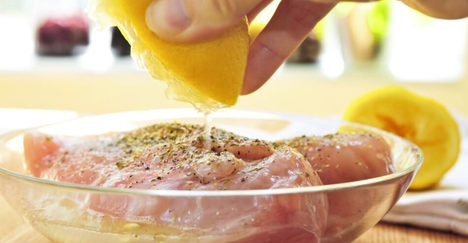 Frango cru em tigela de vidro sendo marinado com ervas, limão e azeite.