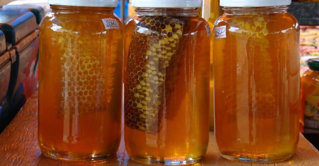 três potes de mel puro de abelha contendo a fava dispostos em uma mesa rustica de madeira.
