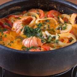 Moqueca de lagosta em uma panela de barro, com pedaços de lagosta, tomate, cebola e pimentão, decorada com coentro fresco.