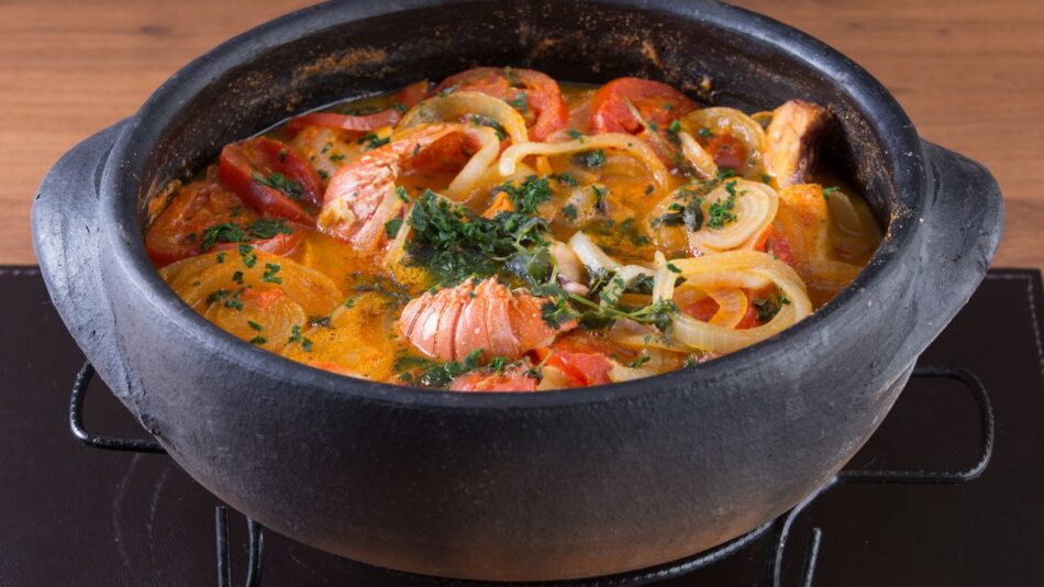 Moqueca de lagosta em uma panela de barro, com pedaços de lagosta, tomate, cebola e pimentão, decorada com coentro fresco.