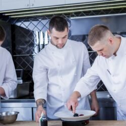 três chefes de cozinha trabalhando em uma bancada de cozinha profissional fazendo a finalização de pratos. O que é gastronomia.