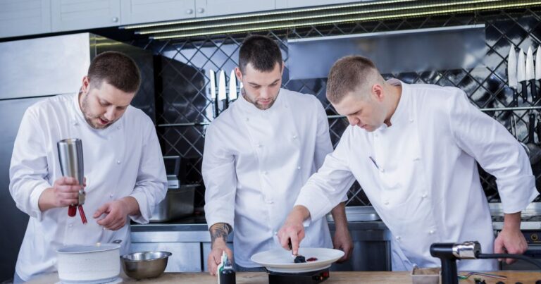 três chefes de cozinha trabalhando em uma bancada de cozinha profissional fazendo a finalização de pratos. O que é gastronomia.