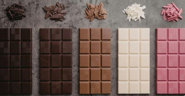 Uma imagem em plano geral exibe cinco barras de chocolate retangulares dispostas horizontalmente e com textura geométrica uniforme de quadrados sobre uma superfície cinza escuro de pedra. Chocolate amargo, meio amargo, ao leite, branco e ruby.