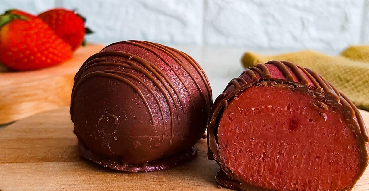 Trufa de morango cortada ao meio, exibindo um recheio intenso e apetitoso com uma casquinha fina de chocolate nobre.