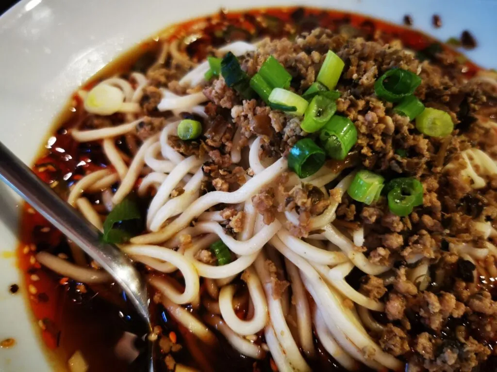 Tigela de Dan Dan Noodles, com macarrão coberto por molho picante, carne moída, amendoim, e cebolinhas, disposta em um prato fundo.