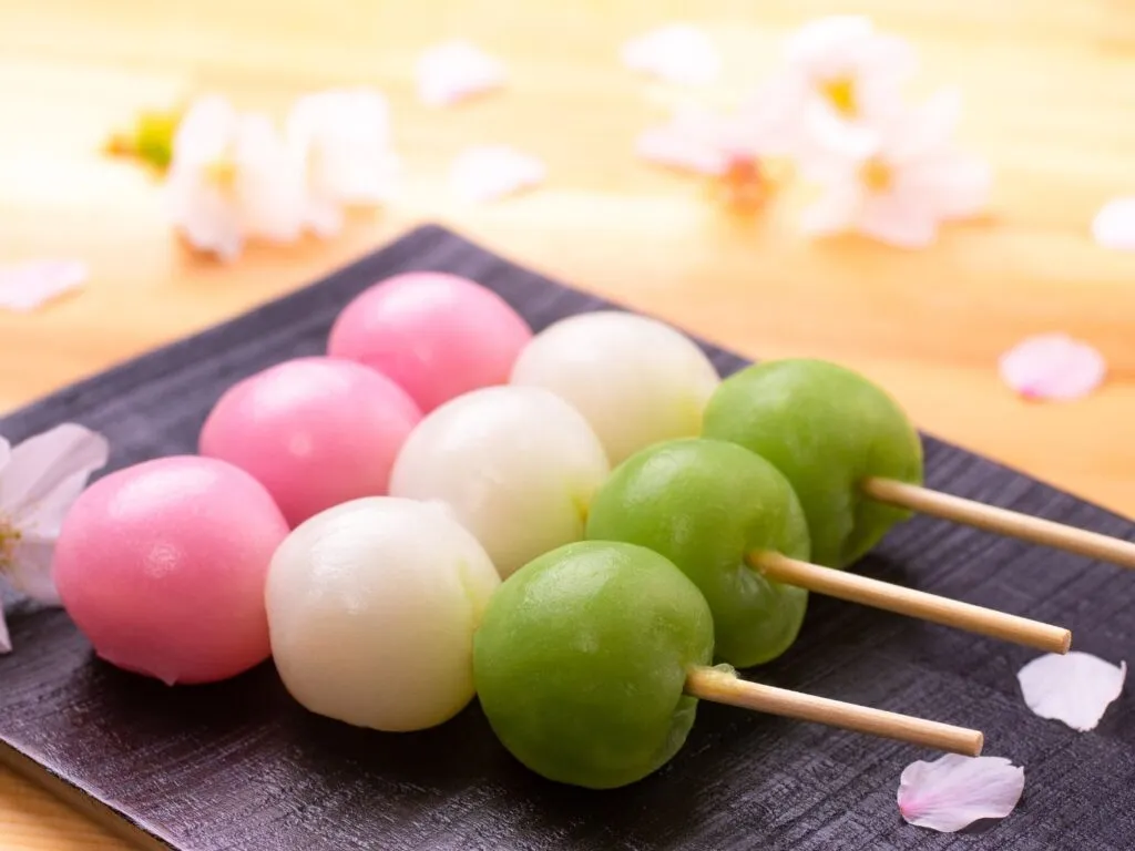 Três bolinhos de dango coloridos, enfileirados em um espeto de bambu, dispostos sobre um prato de cerâmica.