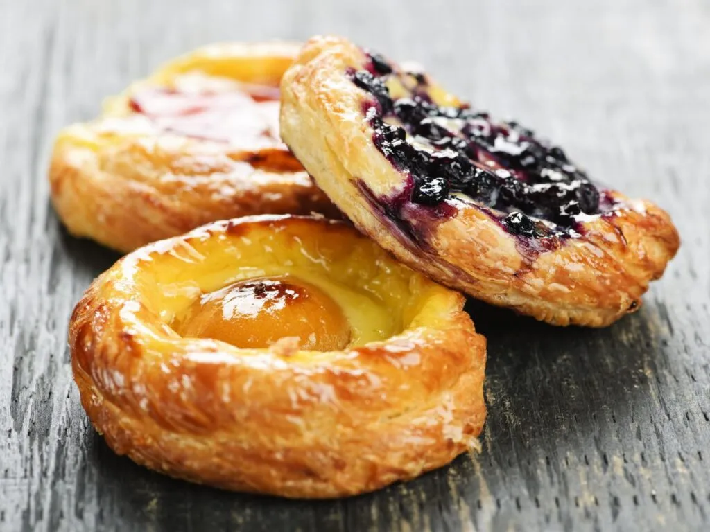 Danish pastry dourada, com camadas folhadas e recheio de creme, frutas e glacê, disposta em um prato de porcelana.