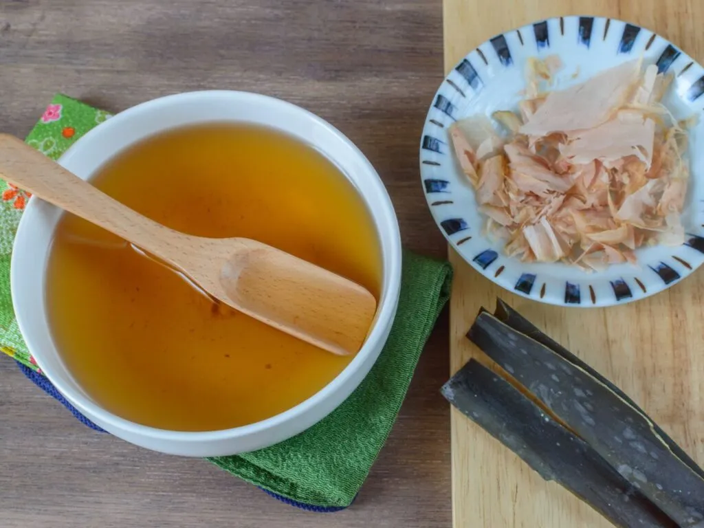 Caldo dashi claro em uma tigela de cerâmica, com algas kombu e flocos de bonito visíveis, sobre uma mesa de madeira.