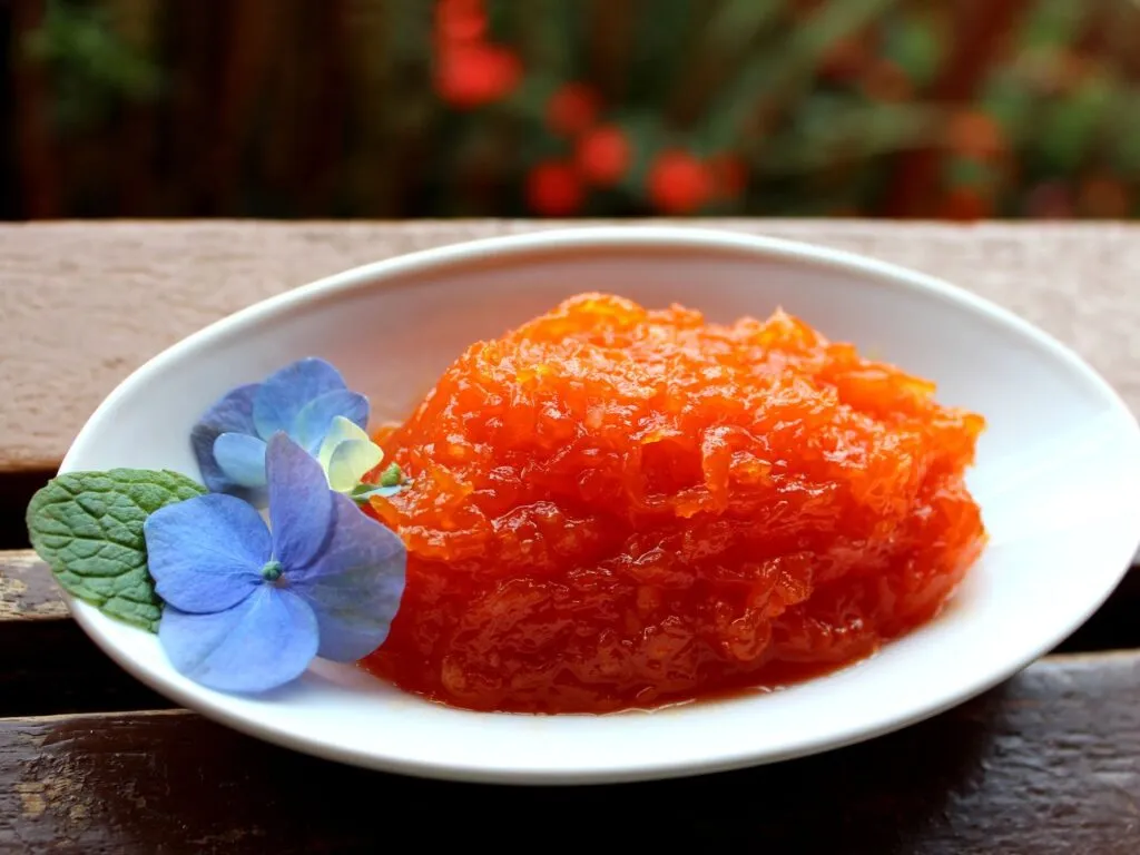 Doce de abóbora ralado em um prato raso, com uma textura granulada e cor laranja vibrante, decorado com canela em pó e flor comestível.