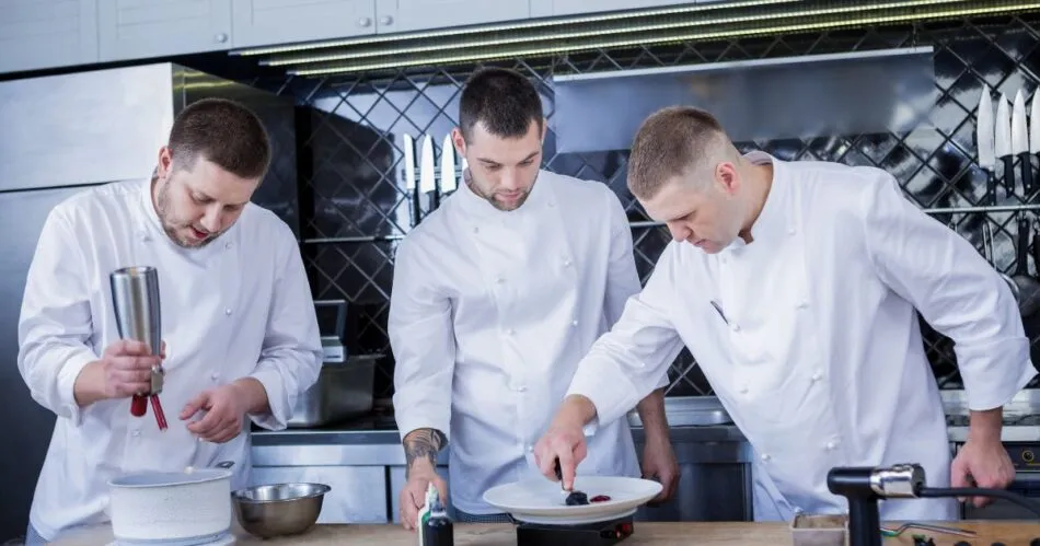 três chefes de cozinha trabalhando em uma bancada de cozinha profissional fazendo a finalização de pratos. O que é gastronomia.